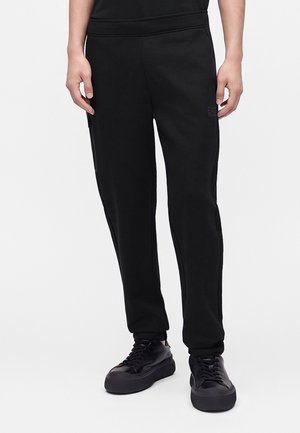 Mand iført sorte slim-fit joggerbukser og sorte lave sneakers mod en ensartet lys baggrund.