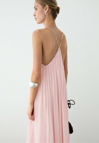 Woman in a light pink pleated dress with thin straps and an open back, wearing a silver bracelet and holding a black bag.