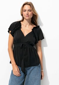 Haut noir avec des manches volantes, design en V et taille cintrée. Le tissu est doux avec une silhouette fluide. Porté avec un pantalon en denim bleu.