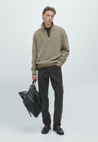 Beżowy sweter dzianinowy z zapięciem na zamek, połączony z ciemnymi dżinsami. Model trzyma czarną skórzaną torbę i ma na sobie ciemne buty.