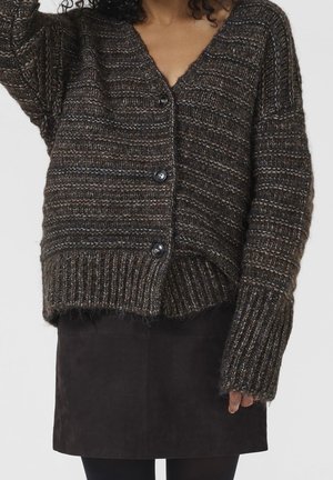 Femme portant un cardigan à gros tricot marron foncé avec de gros boutons et une jupe en suède noire, sur un fond uni.