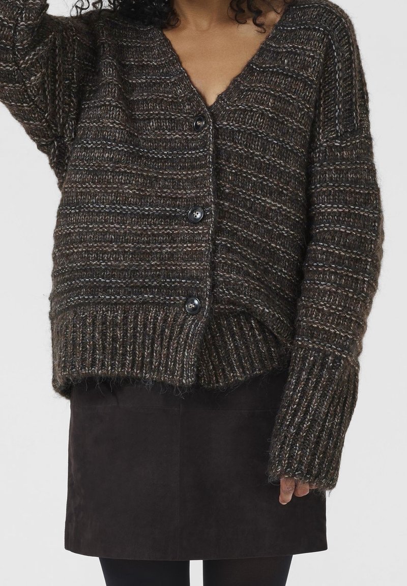 Femme portant un cardigan à gros tricot marron foncé avec de gros boutons et une jupe en suède noire, sur un fond uni.
