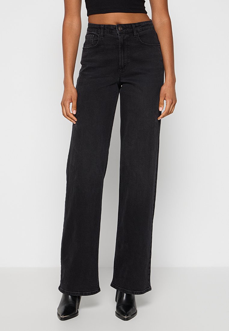 Kvinna som bär högmidjade svarta vida jeans med framfickor och svarta spetsiga anklar stövlar mot vit bakgrund.