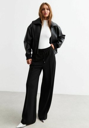 Schwarze Kunstleder-Cropped-Jacke mit breitem Kragen, kombiniert mit hoch taillierten schwarzen Hosen und silbernen Spitzenabsatzschuhen. Glatte Texturen.