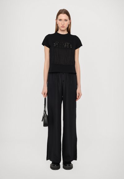 Black short-sleeve t-shirt with fringe detail and decorative elements on the front, paired with loose black trousers and chunky black shoes.