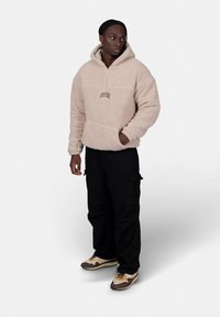Beige fleece pullover hoodie with a half zipper and "CRYSTAL" logo, paired with black cargo pants and brown sneakers with cream accents.