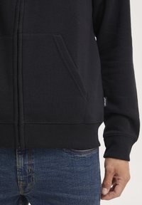 Svart zip-up hettegenser med frontlomme, laget av mykt stoff, med ribbestrikkede mansjetter og kant. Akkompagnert av mørkeblå denimjeans.