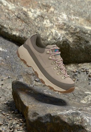 Chaussure de randonnée marron clair avec œillets lacés roses, semelle robuste couleur tan, et logo Columbia, placée sur un terrain rocailleux extérieur.