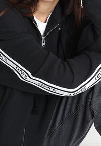 Sudadera con cremallera negra hecha de una mezcla de algodón, con rayas de logo blanco de Levi's a lo largo de las mangas y una cinturilla elástica para un ajuste cómodo.