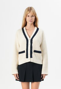 Femeie îmbrăcând un cardigan tricotat crem cu detalii și butoni bleumarin, asortat cu o fustă plisată neagră, stând în fața unui fundal alb.