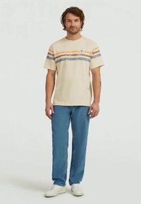 Mann iført kremfarget t-skjorte med oransje, rød og blå horisontale striper, blå jeans og hvite joggesko, stående mot en ensartet bakgrunn.