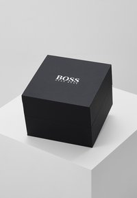 Boîte carrée noire avec une finition mate, arborant un logo "BOSS" en relief blanc sur le dessus. Pas de matériel ou d'accents visibles.