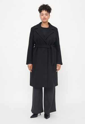 Donna con capelli ricci che indossa un cappotto nero con cintura, pantaloni grigi scuri a gamba larga e décolleté neri a punta, in piedi contro uno sfondo bianco.