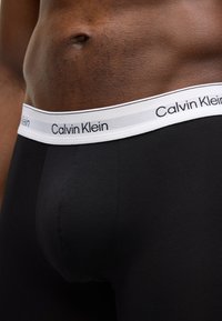 Boxeri negri din bumbac cu talie albă pe care este scris logo-ul „Calvin Klein”. Materialul are o textură netedă și un design fixat.
