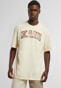 Light yellow oversized t-shirt with short sleeves, featuring brown text "KANI" and "Originato" in a casual font. Soft cotton material.