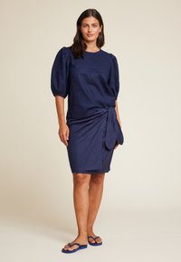Mørkeblå kjole med puffede ermer, omslagdesign og aksent med sidesløyfe. Laget av lett stoff, knelengde med en glatt tekstur.