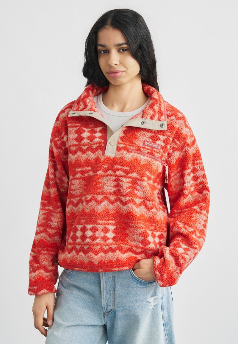 Αντιανεμικό φούτερ από φλις με κόκκινο και λευκό μοτίβο, μισό φερμουάρ, μπεζ γιακά και τσέπη στο στήθος. Φορεμένο με ανοιχτόχρωμο μπλε τζιν.