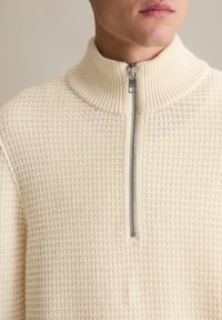 Pullover mit Viertelreißverschluss in Creme, mit strukturiertem Waffelstrickmuster. Gerippter Kragen und silberner Reißverschlussakzent.