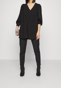 Blusa nera con maniche a sbuffo, con pieghe e bottoni lungo la parte anteriore, abbinata a pantaloni neri aderenti e stivaletti. Borsa a tracolla multicolore.