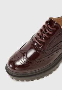 Bordeaux lackskinns brogue skor med perforerade detaljer, rund tå och kontrasterande svart kraftig gummisula. Bruna snöre detaljer.