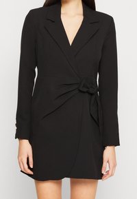 Vestido negro envolvente de tejido suave, con mangas largas, escote en V profundo y un detalle anudado en la cintura, con un corte ajustado.