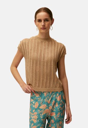 Mujer con un top beige de punto de manga corta y pantalones turquesa con estampado floral melocotón, de pie con una mano en la cadera frente a un fondo liso.