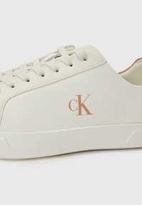Sneaker bianco realizzato in pelle liscia con punta testurizzata, caratterizzato da lacci beige chiaro e un logo di colore rame sottile. Design con suola piatta.