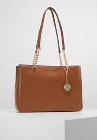 Bruine leren tote bag met dubbele schouderbanden, gouden kettingaccenten en een ronde bedel met het DKNY-logo. Getextureerde afwerking.