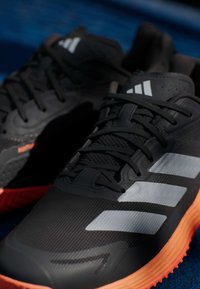 Μαύρα αθλητικά παπούτσια με υφή στην επάνω πλευρά και πορτοκαλί λάστιχο στη σόλα. Διαθέτουν τρεις λευκές ρίγες, στρογγυλή μύτη και μαύρη δαντέλα.