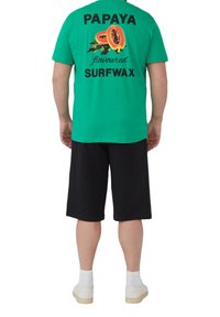 Groene katoenen t-shirt met de opdruk "PAPAYA Flavored SURFWAX" en papaya afbeeldingen op de achterkant, gecombineerd met zwarte shorts en witte sokken.