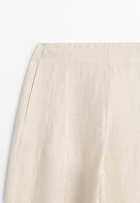Béžové lněné kalhoty s vysokým pasem, jemnými záhyby a skrytou boční kapsou, zobrazené od pasu po polovinu stehna.