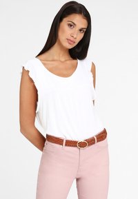 Witte mouwloze top met ruches op de schouders, gecombineerd met roze jeans en een gevlochten bruine riem met een ronde gesp.