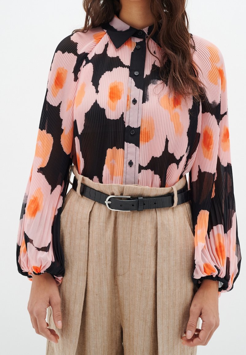 Långärmad blus med ett svart och rosa blommönster, strukturerade plisseringar och en svart knäppkrage, tillsammans med högmidjade beige byxor och ett svart bälte.