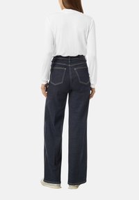 Jeans z wysokim stanem w ciemnym deninie, o szerokim kroju nogawki, widocznych tylnych kieszeniach i kontrastowym białym przeszyciu. W połączeniu z białą bluzką z długim rękawem.