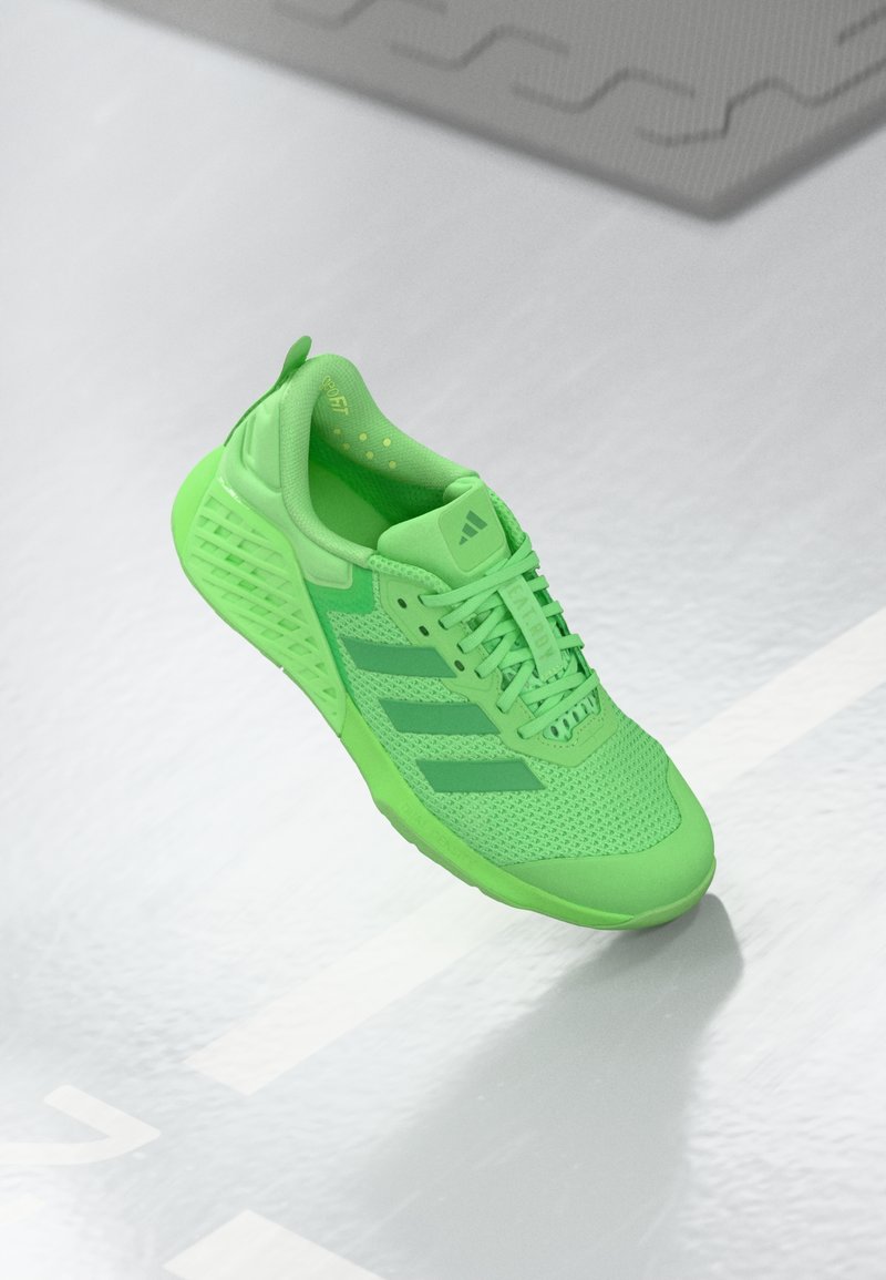 Chaussure de sport verte avec dessus en mesh, détails texturés et trois bandes vertes plus foncées. Dispose d'une semelle épaisse et rembourrée pour un bon soutien.