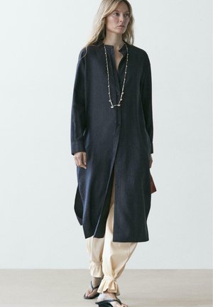 Donna con cappotto nero lungo abbottonato, pantaloni beige aderenti, sandali neri e una lunga collana di perline che cammina su uno sfondo bianco uniforme.