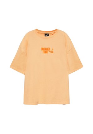 Jeune homme portant un t-shirt graphique orange, un short à carreaux verts, des baskets beiges et un pull bleu drapé sur les épaules sur fond blanc.