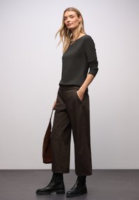 Dunkelgrünes Langarmshirt, braune, gestreifte, weitgeschnittene Hose mit Taschen, schwarze Knöchelstiefel und eine braune Schultertasche vor hellem Hintergrund.