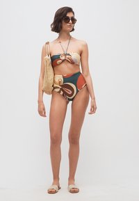 Γυναίκα που στέκεται, φοράει πολύχρωμο μπάντεου μπικίνι με σχέδιο, σκούρα γυαλιά ηλίου, πλεκτή τσάντα ώμου και σανδάλια με ανοιχτή μύτη, σε λευκό φόντο.