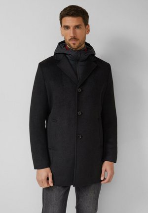 Mężczyzna w czarnym, zapinanym na guziki płaszczu na kurtkę z kapturem, stojący na tle jednolitego szarego tła.