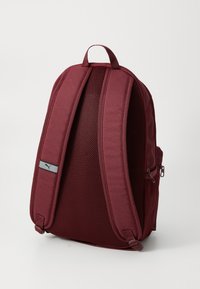 Maroon ruksak od tkanine s prozračnom leđa. Ima podstavljene trake i mali sivi logotip u blizini vrha.