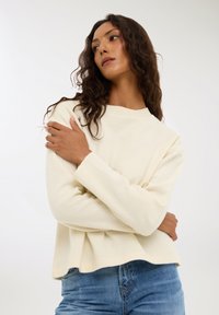 Sweter w kolorze kremowym z żebrowanym okrągłym dekoltem i długimi rękawami, zestawiony z niebieskimi dżinsami. Miękka, teksturowana tkanina, luźny krój.