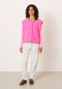 Svetlo roza brezrokavnik iz flisa z mehkobno teksturo in zaobljenim ovratnikom. Ima gumbe spredaj in majhen logotip na označbi. Kombiniran s svetlimi kavbojkami in rjavimi čevlji.