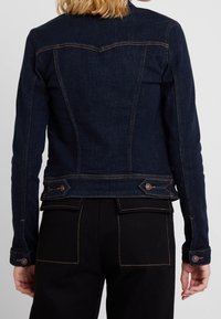 Mørkeblå denimjakke med en tætsiddende design, der har fremtrædende syninger, knapdetaljer og en struktureret ryg. Detaljerede lommer er synlige.