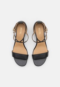 Sandales en cuir noir avec une large bride au niveau des orteils, des brides étroites à la cheville et une semelle intérieure beige. Présente des détails en métal doré.