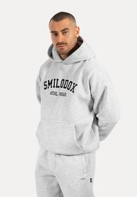 Hoodie gris en tissu doux, avec une poche kangourou à l'avant et le texte noir "SMILODOX ATHL. WEAR" sur la poitrine.