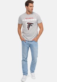 Šedé bavlněné tričko s nápisem a logem "ATLANTA FALCONS" v černé a červené barvě, spárované s světle modrými džíny a bílými teniskami.