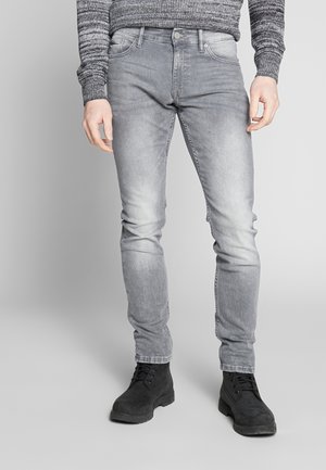 Mand iført tætsiddende lysegrå cowboybukser, sorte snørebåndsstøvler og en grå strikket sweater mod en ensfarvet baggrund.