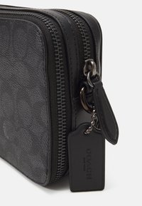 Bolso de cuero negro con cremallera, presenta un patrón texturizado, herrajes plateados y una pequeña etiqueta de identificación unida por una cadena.