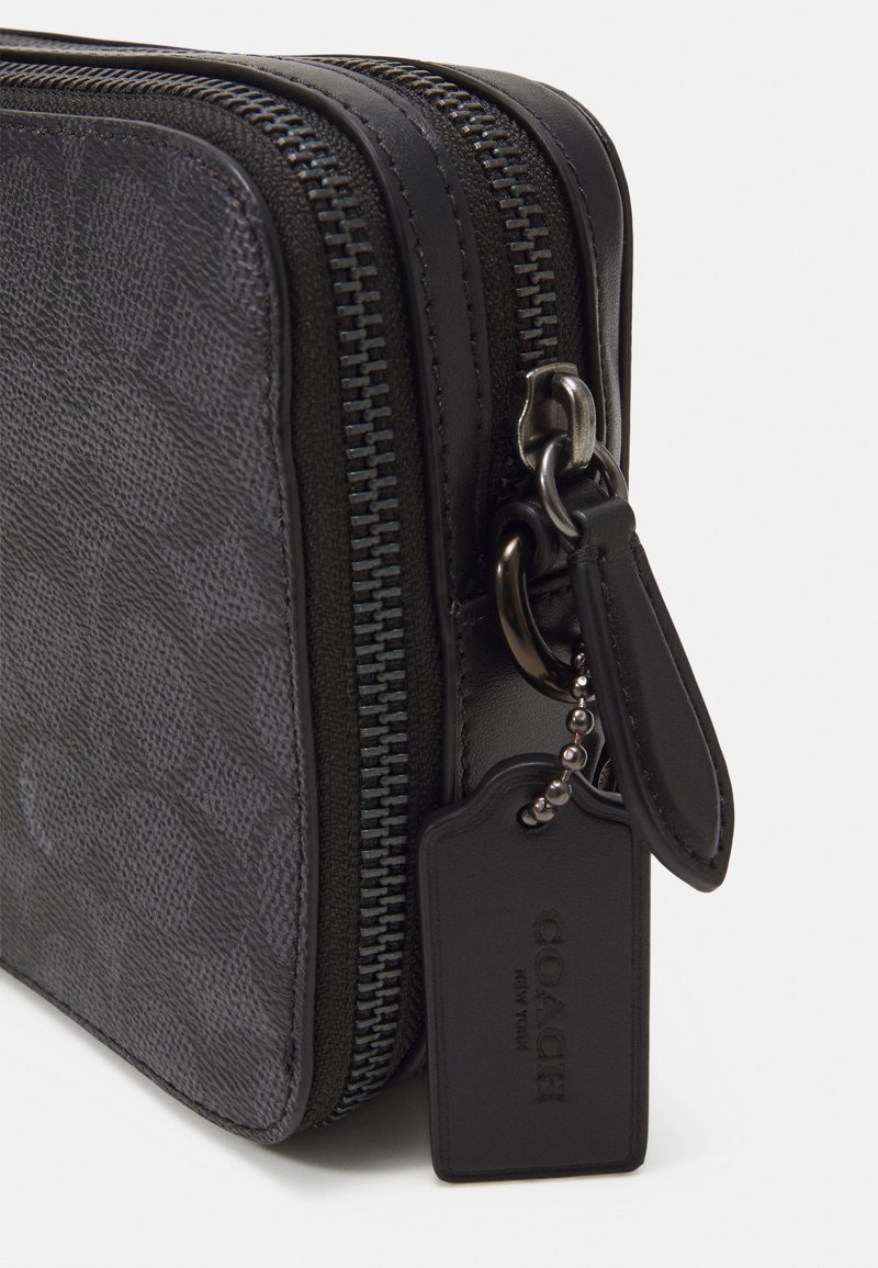 Bolso de cuero negro con cremallera, presenta un patrón texturizado, herrajes plateados y una pequeña etiqueta de identificación unida por una cadena.